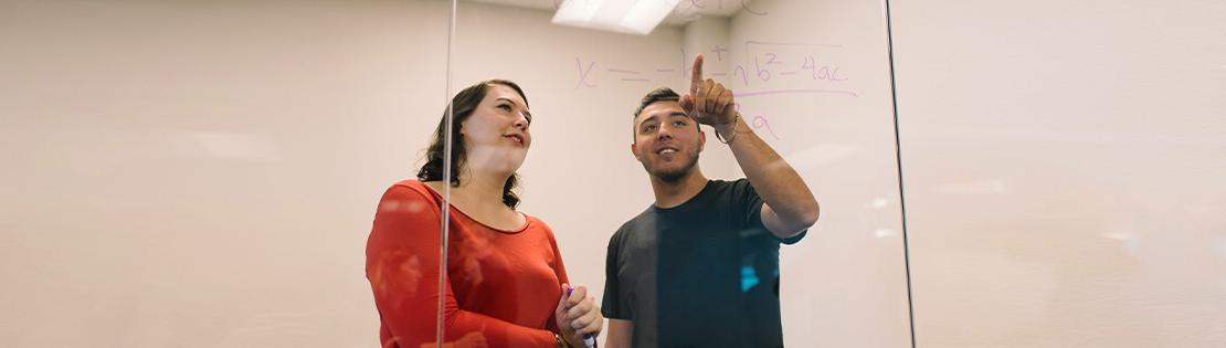 Two Pima students work on a math problem together on a window in a Pima study room